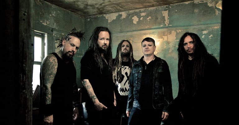 Five band members in dark clothing stand against a weathered turquoise wall in a gritty indoor setting.