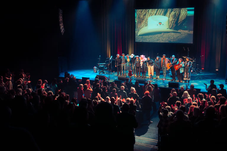 Large audience watches performers on stage beneath a projection screen in a theatre lit by blue spotlights.