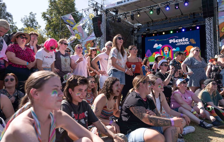 Crowd of festival-goers seated on grass watching performers on an outdoor stage lit by purple spotlights.