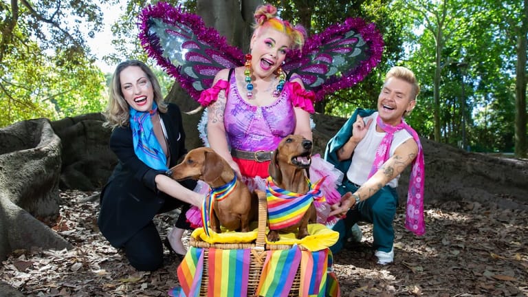 Three performers in colourful costumes hold a striped prop basket outdoors amongst trees and purple foliage.