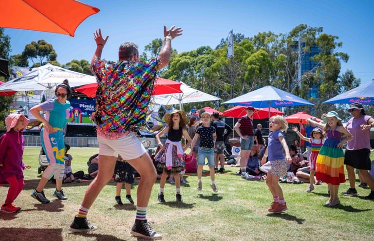 Children and families dance and play on grass at an outdoor community festival under red and white marquees.