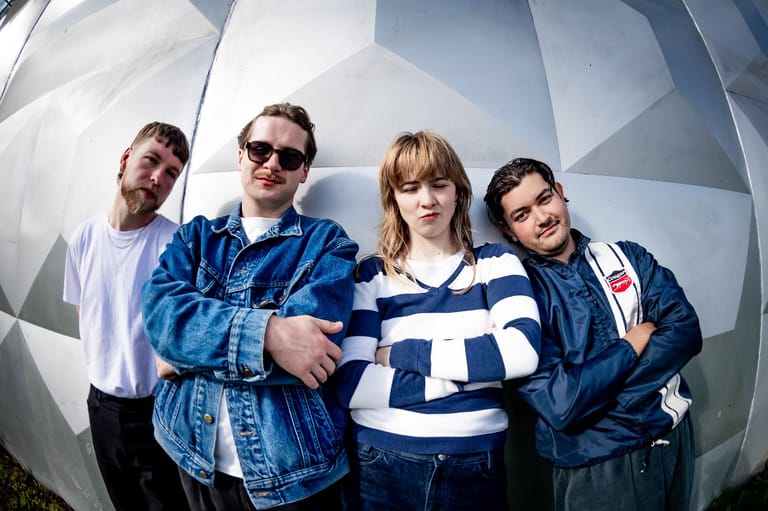 Four band members stand together against a large white geometric backdrop, dressed in casual streetwear and sunglasses.