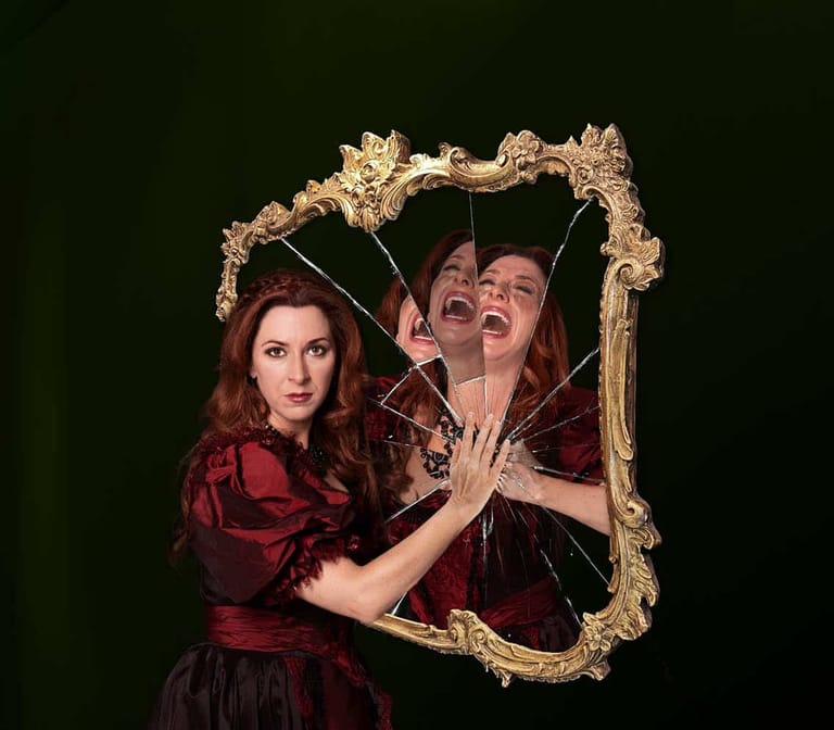 Two performers in burgundy costume pose with an ornate gold-framed mirror against a dark green background.