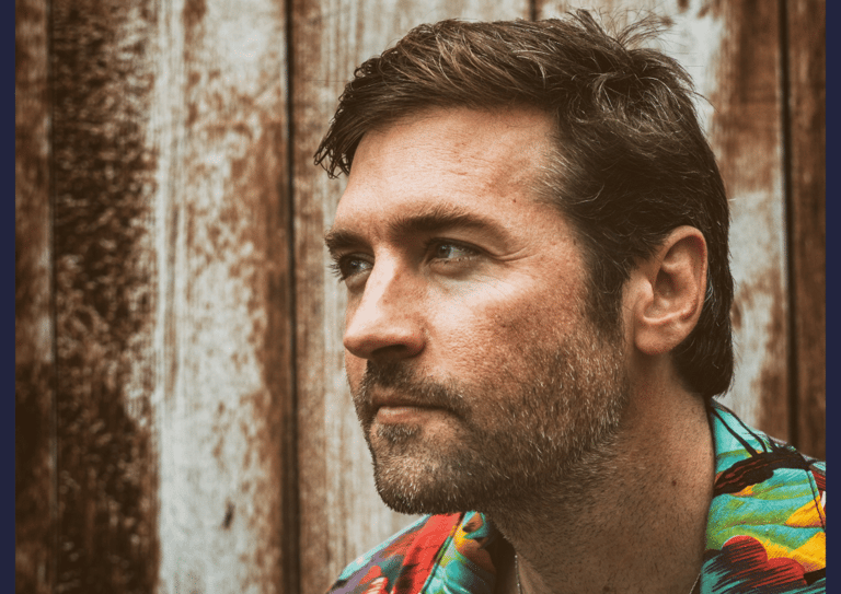 Man with dark hair and beard wearing a colourful striped shirt, photographed in profile against a weathered corrugated metal background.