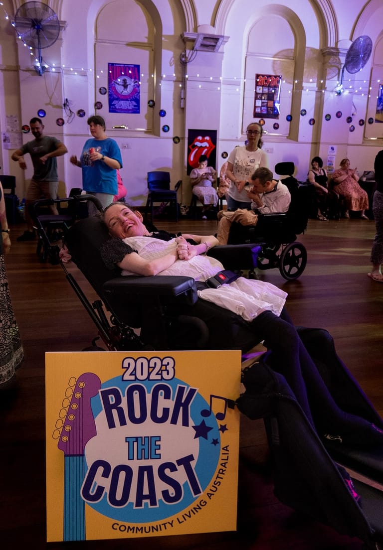 Audience members in wheelchairs and seated positions enjoy Rock the Coast community event in a purple-lit venue with ornate arches.