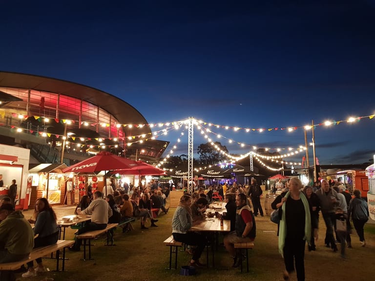 Outdoor night market with illuminated red pavilion, string lights and crowds browsing vendor stalls at dusk.