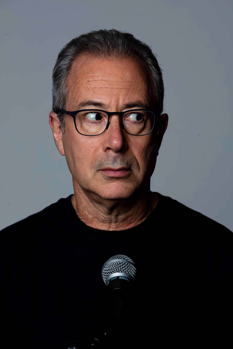 Man wearing glasses and black shirt holding a microphone against a grey background.