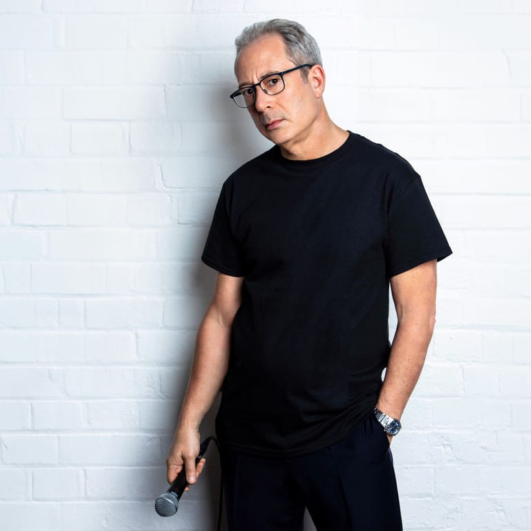 Man in black t-shirt and glasses standing against white brick wall.