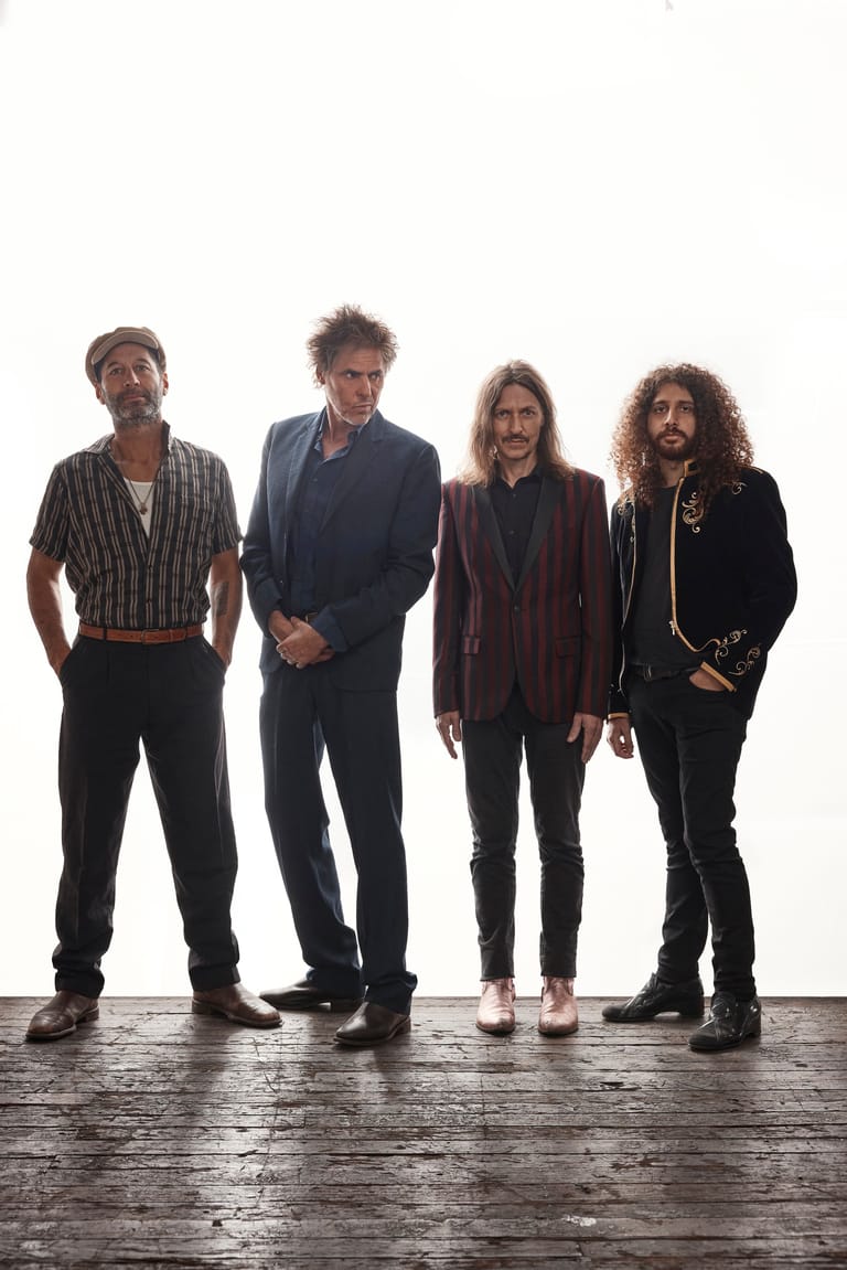 Four band members stand in a line against a white backdrop, dressed in dark casual and smart-casual clothing.