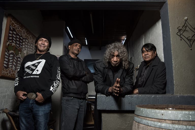 Four musicians in black jackets stand together in a dimly lit industrial space with a barrel and brick wall visible.