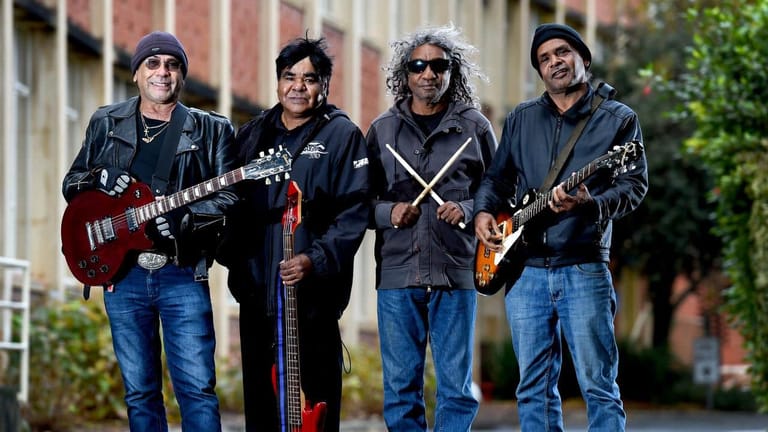 Four musicians in dark jackets and jeans pose with guitars outside a brick building, holding their instruments in a band formation.