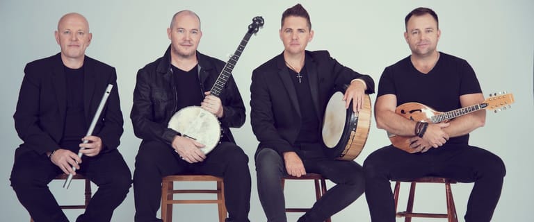 Four musicians in black clothing sit holding acoustic guitars and percussion instruments against a white background.