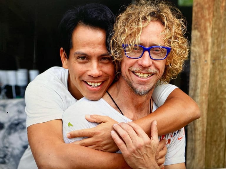 Two men smiling at the camera, one wearing glasses and embracing the other from behind outdoors.