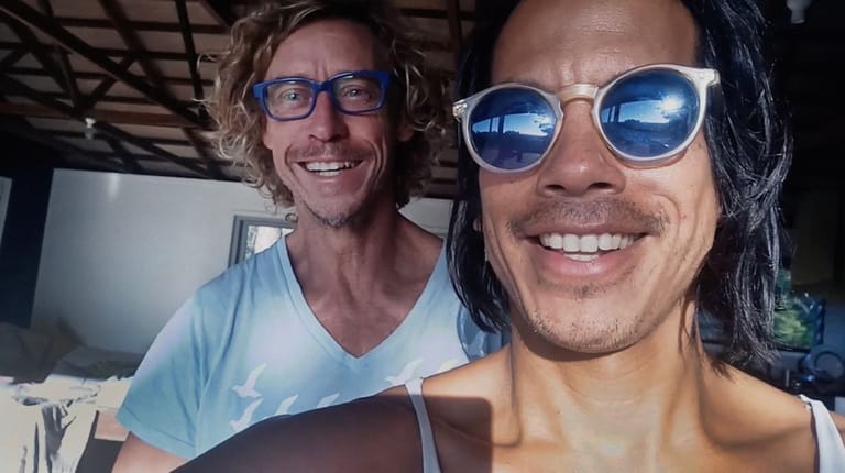 Two smiling men pose for a selfie indoors, one wearing glasses and a light blue shirt, the other in reflective sunglasses.