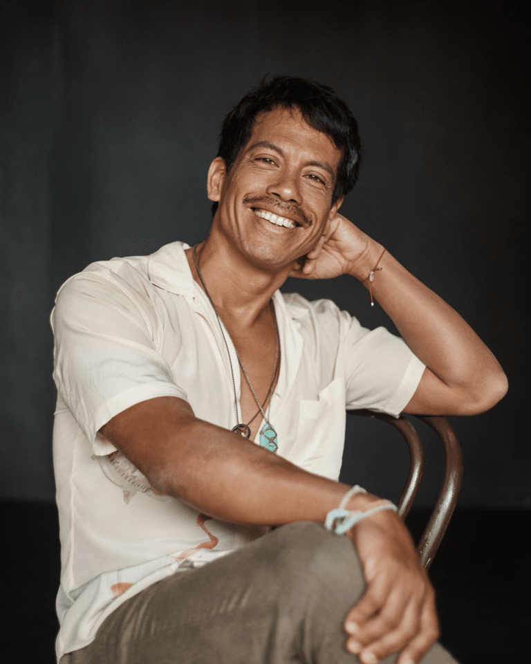 Man in white shirt smiling at camera, resting head on hand against dark background.