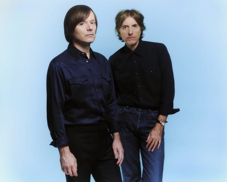 Two musicians stand against a light blue background, one in a dark navy shirt and the other in a black top.
