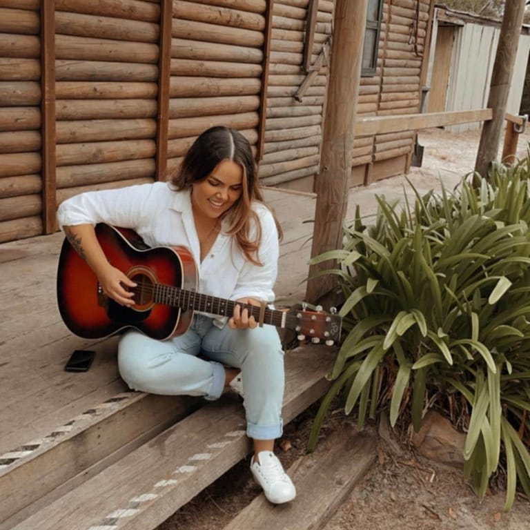 Musician crouched outdoors playing acoustic guitar beside a potted plant against wooden fence panelling.
