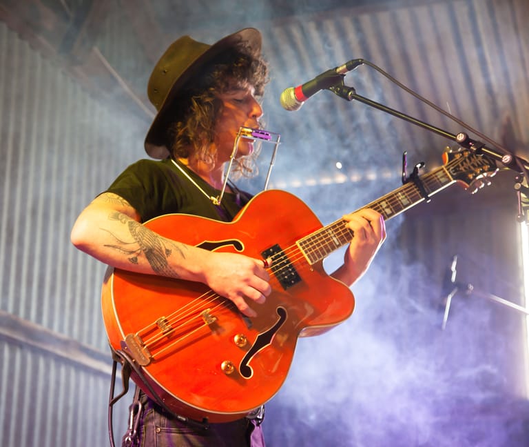 Musician in a brown hat playing a red semi-acoustic guitar at a microphone under purple stage lighting.