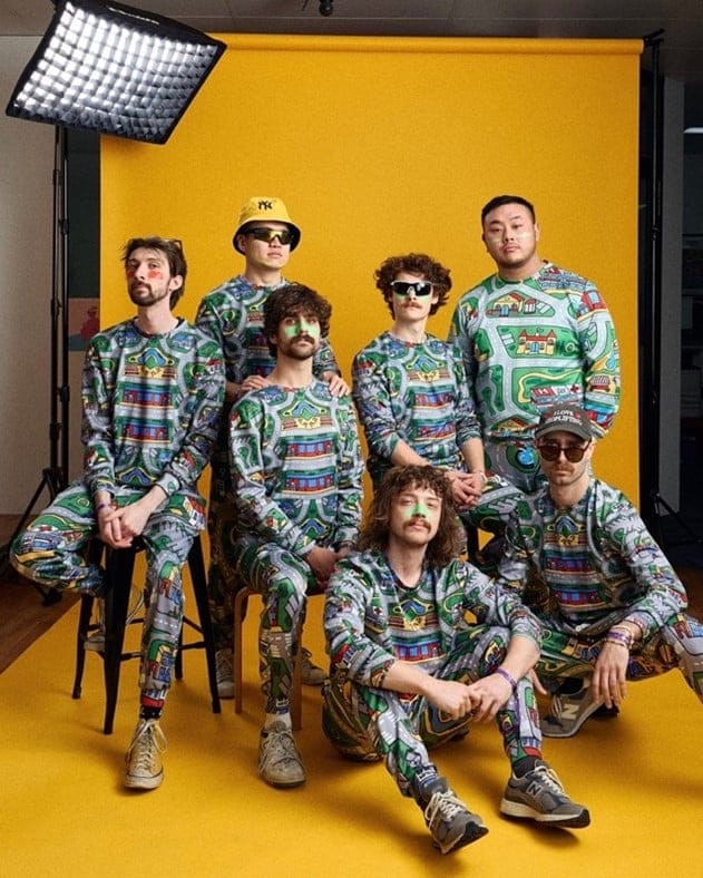 Seven band members in matching patterned jumpsuits pose against a bright yellow studio backdrop under stage lighting.