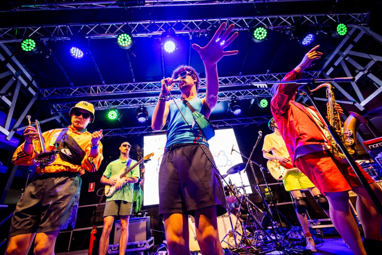Five-piece band performing on stage under blue and purple stage lighting with a white screen backdrop.