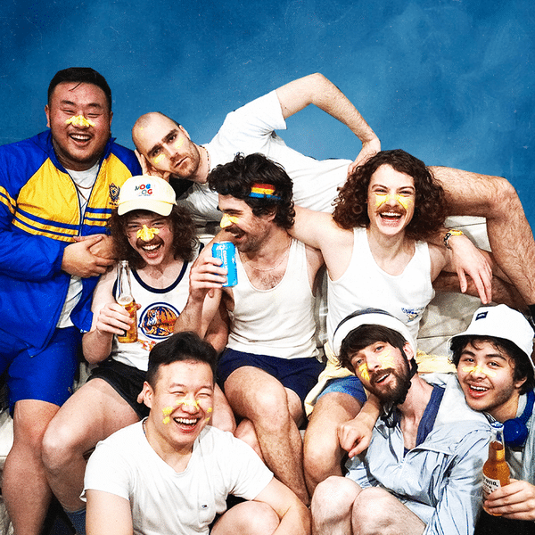 Group of seven smiling men posing together against a blue backdrop in casual summer attire.