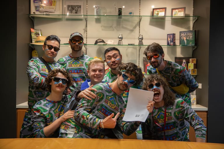 Group of people in matching patterned shirts gathered around a laptop, posing together in front of glass display cabinets.
