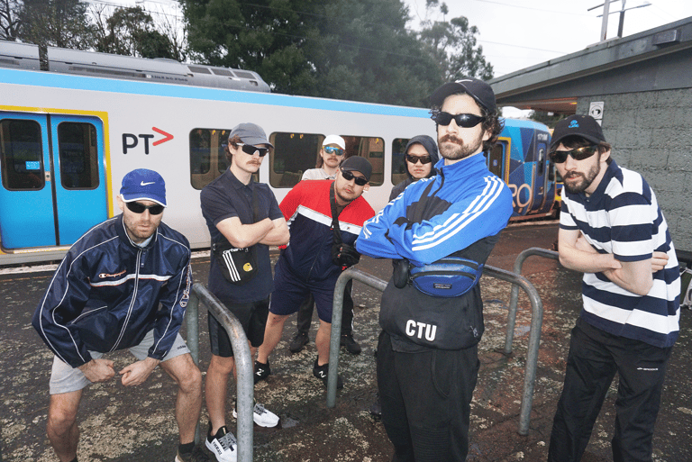 Six people in casual attire stand in front of a blue PTV train on muddy ground outdoors.