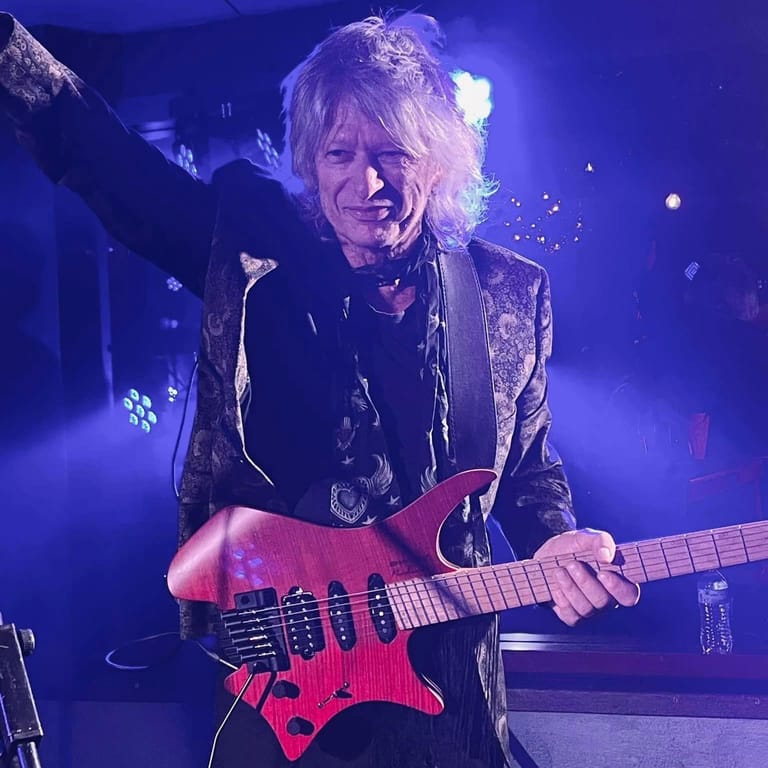Guitarist performing on stage with a red electric guitar, lit by blue and purple stage lighting.