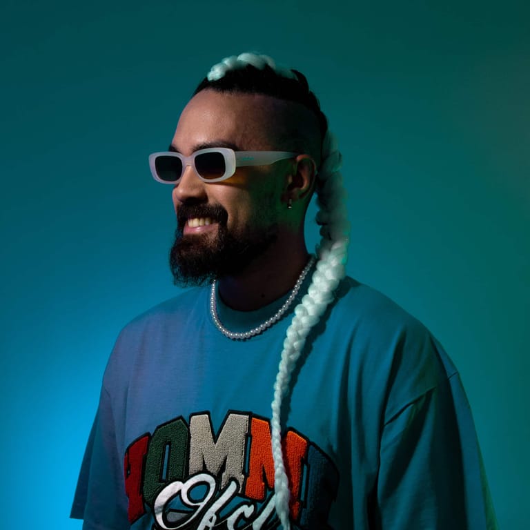Man wearing white sunglasses and pearl chain necklace against teal blue background in studio portrait.