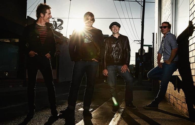 Five musicians stand in a sunlit warehouse space with dramatic shadows cast across the floor.