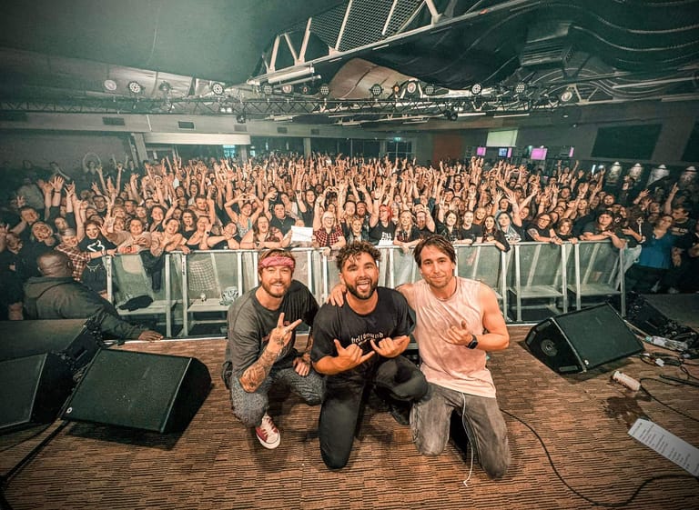 Three men crouch on stage before a packed crowd in a large indoor venue with industrial ceiling fixtures.