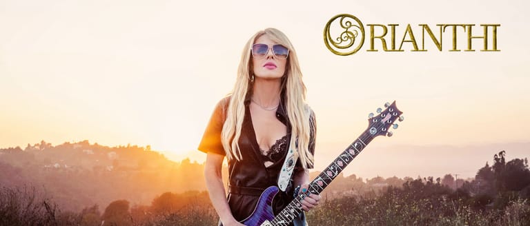 Musician with long blonde hair holding a purple guitar at sunset in a rural landscape.