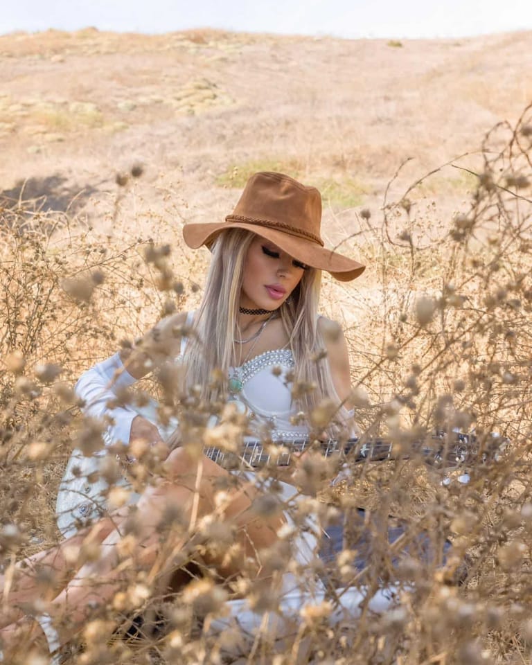 Person in brown cowboy hat and white dress standing among dry cotton plants in arid landscape.