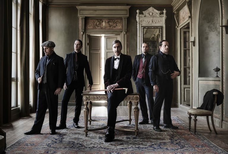 Five men in formal attire stand in an ornate, classical interior with period furnishings and artwork.