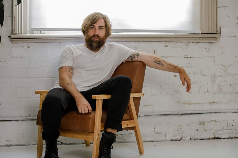 Man with beard seated on wooden chair in white studio space.