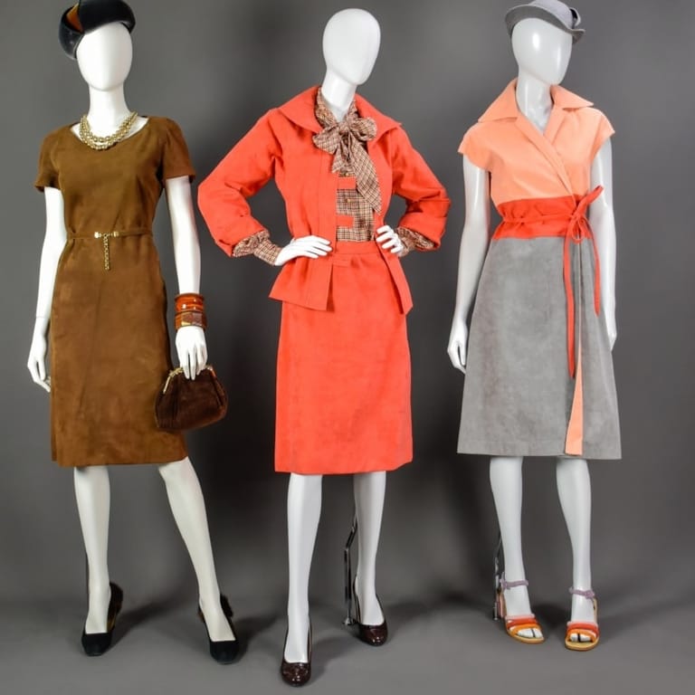 Three mannequins display vintage 1960s dresses in brown, coral and grey with coordinating accessories against a neutral background.