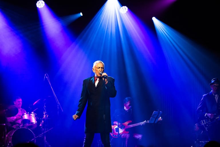 Singer in dark suit performing centre stage under blue and purple spotlights with live band.
