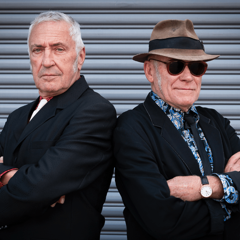 Two men in dark suits stand back-to-back against a corrugated metal wall, one wearing sunglasses and a fedora hat.