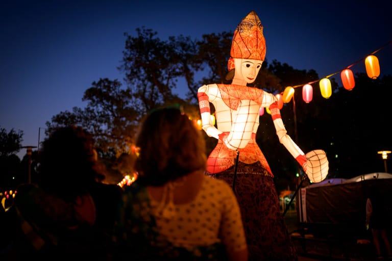 Illuminated figure of a man in orange and white clothing at dusk, with lanterns and spectators gathered nearby.