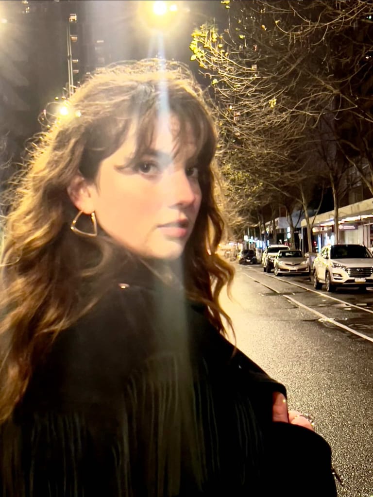 Woman with wavy brown hair and hoop earrings on a night-lit street lined with parked cars.