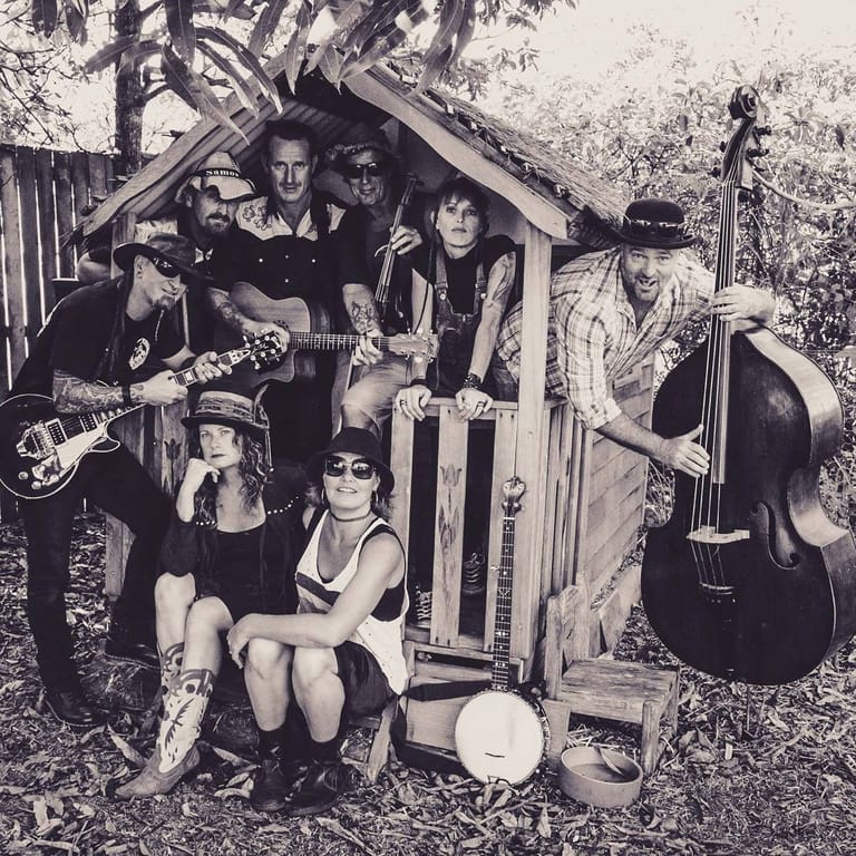 Five musicians pose with guitars and upright bass outside a wooden garden shed in this black and white photograph.