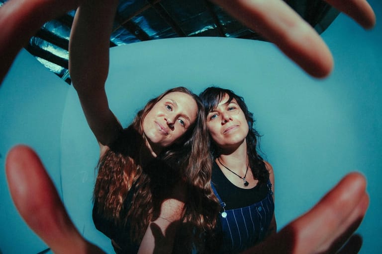 Two performers with long dark hair pose against a bright blue background, framed by hands in the foreground.