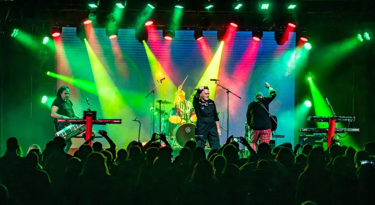 Band performing on stage with vibrant coloured lighting and raised hands in audience.