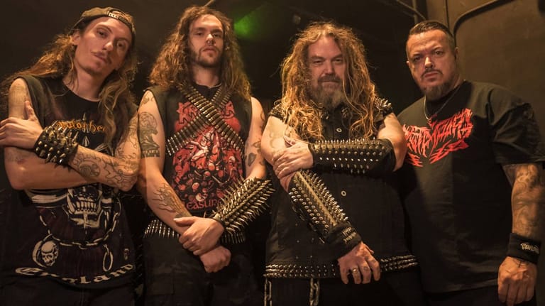 Four heavy metal musicians in studded leather and band shirts stand together on stage under warm lighting.