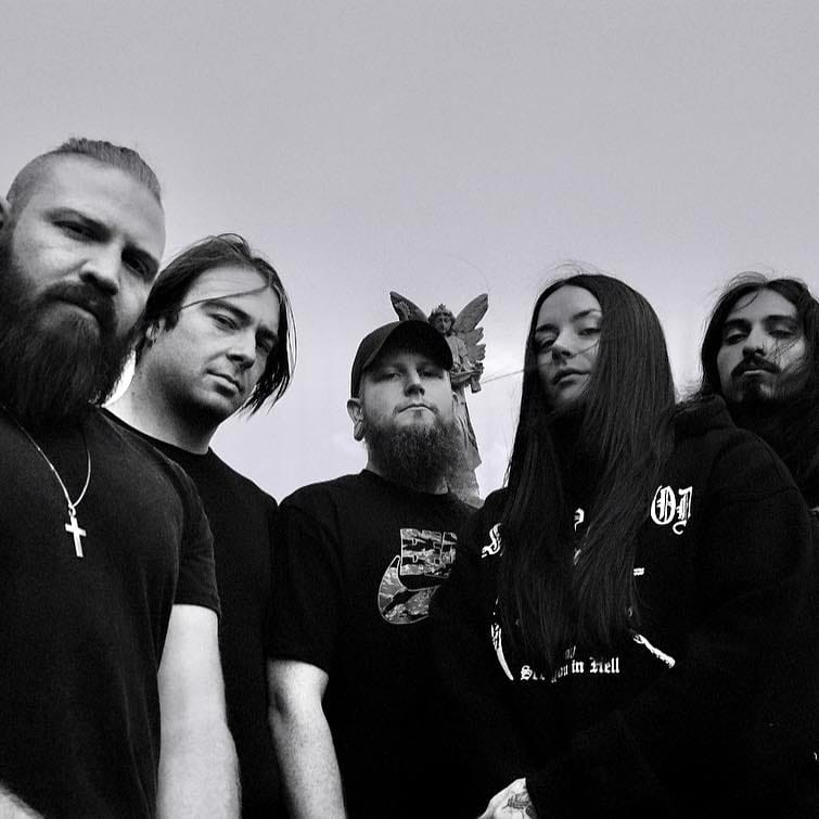 Five band members in black clothing posed against a grey sky in monochrome.