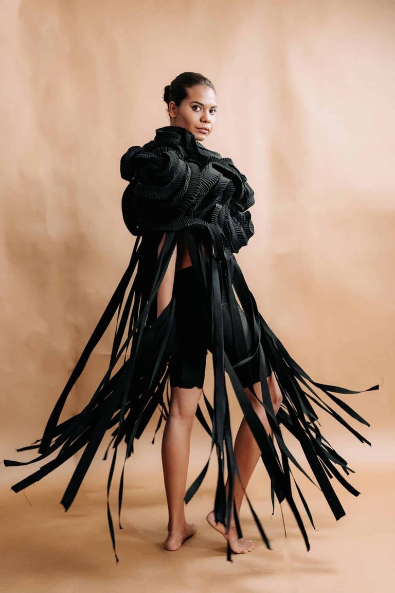 Person wearing an avant-garde black fringed garment against a neutral beige backdrop, gazing over their shoulder.