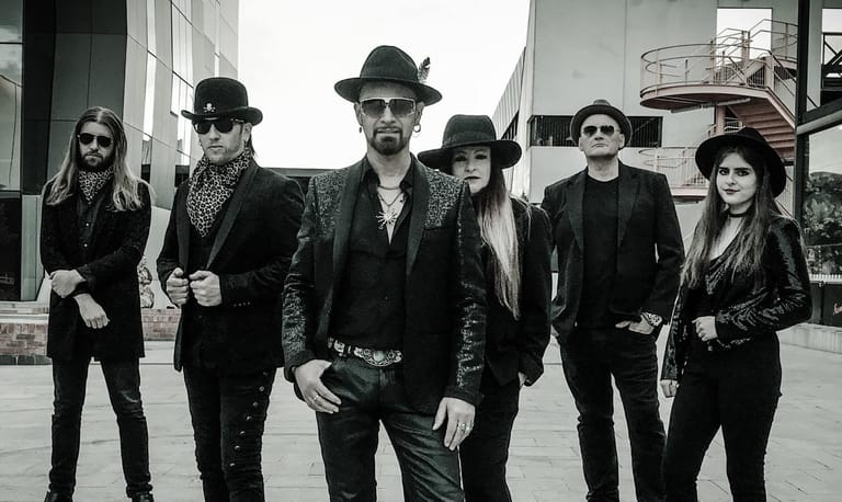 Six performers in black suits and hats pose together on an urban street in a monochrome photograph.