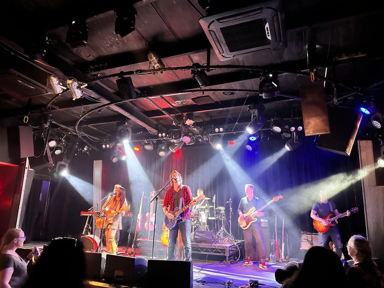 Five musicians perform on a stage bathed in blue and amber stage lighting, with audience members visible in the foreground.