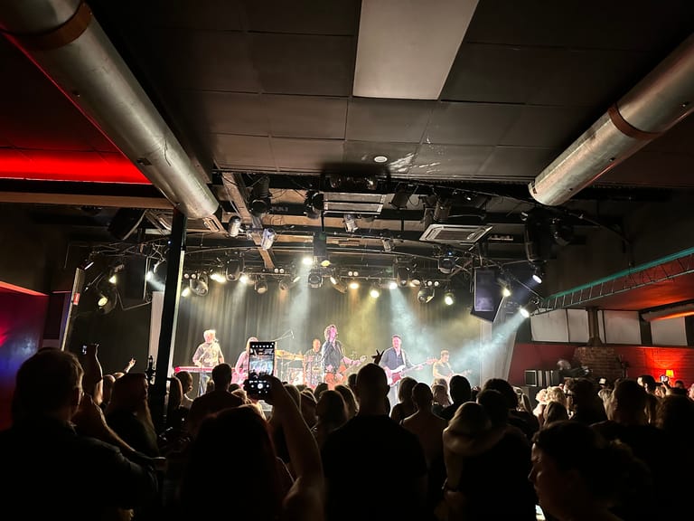 Crowd of concertgoers with raised phones facing a stage lit by spotlights during an indoor live music performance.