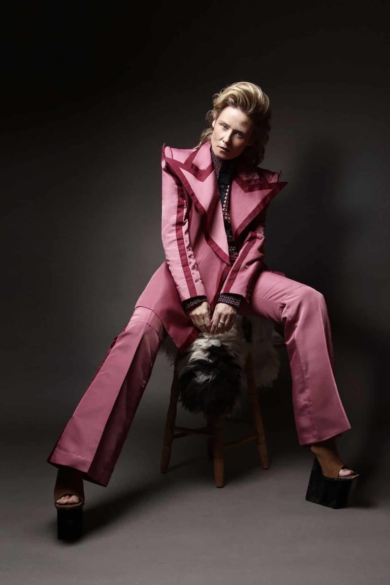 Person in a pink tailored coat sitting on a black stool against a dark background.